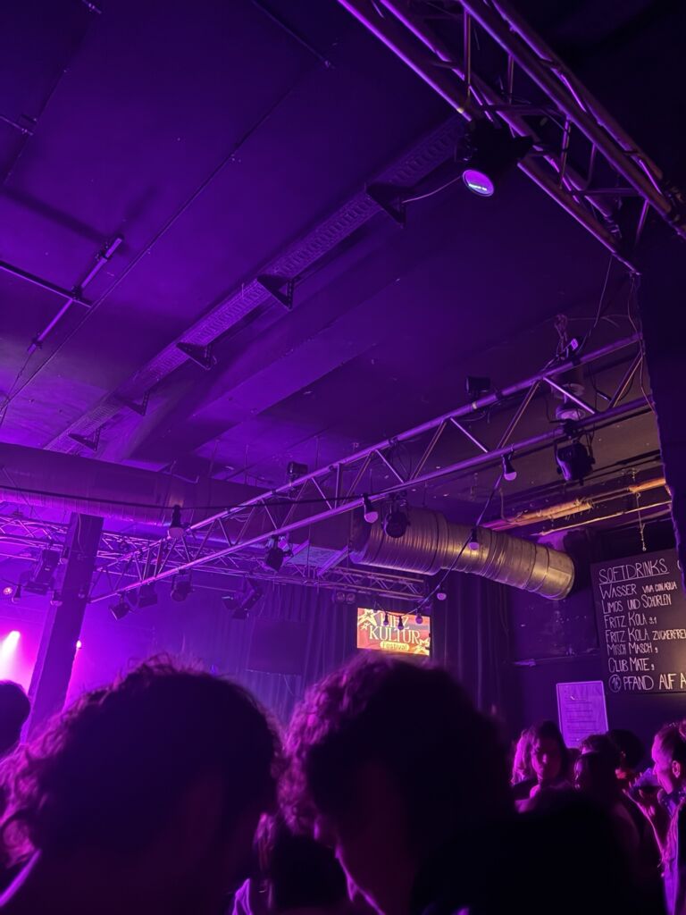 Publikum im violett beleuchteten Konzertsaal mit KiezKultur-Festival-Schild